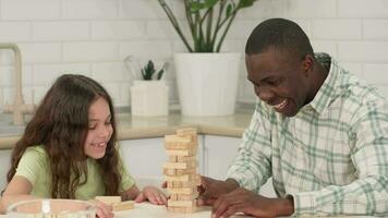 Cheerful African American dad and daughter play board game at home removing wooden blocks from the tower. The tower falls and dad loses. video