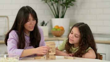 alegre mãe e feliz filha estão jogando uma borda jogos às casa às a mesa removendo de madeira blocos a partir de a torre. video