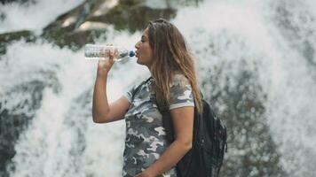 aantrekkelijk meisje drankjes verkoudheid water van een fles en blust dorst terwijl staand Aan een steen in de buurt een waterval. kant visie video