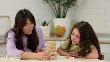 Joyful mother and cheerful daughter are playing a board game at home at the table removing wooden blocks from the tower. The tower falls and mom laughs. video