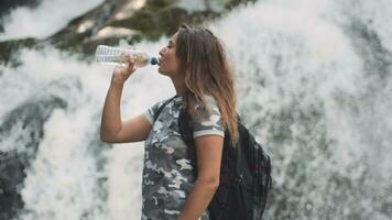 Attractive girl drinks cold water from a bottle and quenches thirst while standing on a stone near a waterfall video