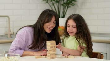 Joyful mother and cheerful daughter are playing a board game at home at the table removing wooden blocks from the tower. video