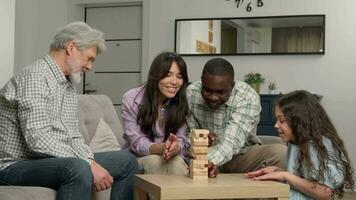 alegre multiétnico familia de diferente años generaciones jugando tablero juego a hogar quitando de madera bloques desde el torre. video