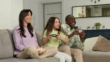 Multi ethnic family playing video game at home sitting on sofa. Daughter wins dad and rejoices.