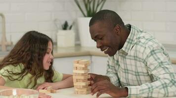 un africano americano papá y hija son jugando un tablero juego a hogar quitando de madera bloques desde un torre. video