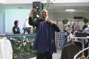 sonriente árabe hombre blogger promoviendo Moda marca mientras grabación vídeo en teléfono inteligente en ropa almacenar. alegre hombre de influencia demostración chaqueta a social medios de comunicación canal seguidores foto