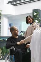 Arab man in wheelchair receiving guidance from asian woman seller while shopping for formal apparel. Boutique customer with disability examining beige jacket while consultant holding hanger photo