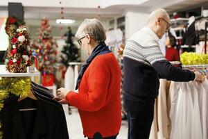 Elderly couple searching for blazers in clothing store with festive decorations, buying items as presents for family on christmas eve. Senior man and woman browsing through merchandise on discount. photo