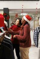 Retail worker showing items to client for shopping assistance during christmas season sales, recommending right clothes size for holiday event preparations. Diverse women discussing fashion products. photo