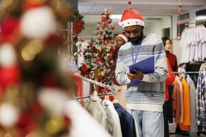 In festive textiles store, sales associate wears santa hat and completes inventory of goods in stock while holding clipboard. Store helper summing every item and arranging clothes. photo