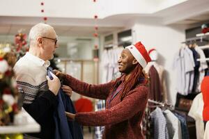 Employee presenting clothes for customer, helping man to choose perfect blazer for festive christmas dinner event. African american store assistant choosing clothing for elderly client. photo
