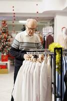 Senior adult looking for clothing items to make christmas present at festive dinner with family, holiday season promotions. Man customer searching for clothes in shopping mall, buying gifts. photo