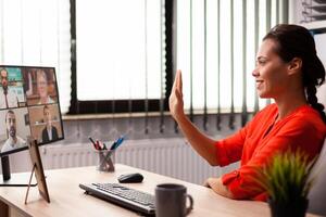 Businesswoman greeting employee with wave during video conference from workplace. Entrepreneur using internet connection for video meeting with coworkers looking at webcam. photo