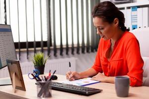 Businesswwoman entrepreneur writing financial sales on charts sitting at desk in workplace wearing red. Successful financial auditor in data marteking looking at graphs on computer screen and writing report. photo