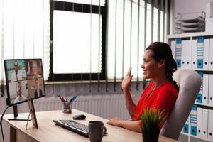 Businesswoman greeting employee with wave during video conference from workplace. Entrepreneur using internet connection for video meeting with coworkers looking at webcam. photo