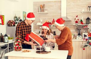 Little girl get surprised with gift boxes for chritmas from senior man and woman wearing santa hat. Happy loving grandparents celebrating winter holidays and relationship with granddaughter in home with x-mas decoration. photo
