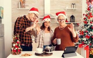 Senior parents and child celebrating christmas and talking with family during online call . Happy multi generation family wearing santa hat during video conference celebrating winter holidays, decorated kitchen. photo