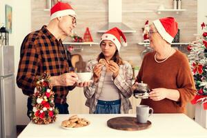 sobrina disfrutando hora con abuelos celebrando navidad y teniendo sabroso galletas para desayuno. contento niño vistiendo Papa Noel sombrero comiendo delicioso galletas desde mayor hombre y mujer mientras celebrando invierno Días festivos con ellos foto