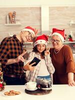 Niece taking selfie with granddparents celebrating christmas in decorated kitchen. Happy cheerful multi generation family using phone to take photo wearing santa hat doing hand gesture with xmastree in the background.