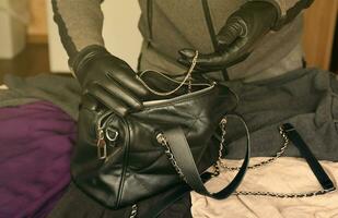 ladrón con traje negro y guantes que se ven en una bolsa de mujer robada abierta. el ladrón saca un collar de oro del bolso de una mujer en la cocina foto