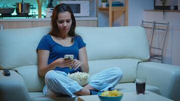 Woman scrolling on phone eating popcorn and watching a movie. Lonely amused happy lady reading, writing, searching, browsing on smartphone laughing amusing using technology internet relaxing at night. photo