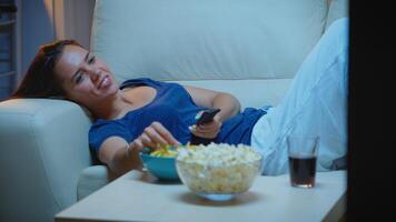 joven mujer en pijamas descansando acostado en sofá en frente televisión sonriente. feliz, divertido, solitario dama disfrutando el noche sentado en cómodo sofá acecho televisión comiendo palomitas de maiz y meriendas foto