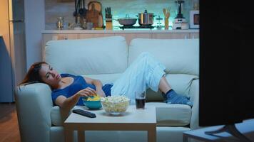 Young woman watching TV and feeling bored sitting on the couch in the living room at home. Tired, exhausted, lonely lady relaxing watching tv lying on comfortable sofa eating snacks late at night photo