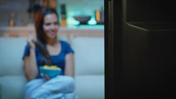 Lady eating snacks while watching tv sitting on sofa. Young happy, excited, amused, home alone lady in pajamas enjoying the evening sitting on comfortable couch eating popcorn in front of television. photo