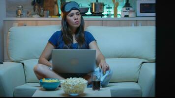 Woman with eye mask using laptop at night while watching tv and eating snacks. Happy person in pijamas sitting on sofa reading writing searching browsing on notebook using internet checking mails photo