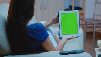 joven dama participación un tableta con verde pantalla sentado en sofá. mujer lectura, mirando a modelo croma llave aislado inteligente cuaderno monitor utilizando tecnología Internet acostado en cómodo sofá. foto