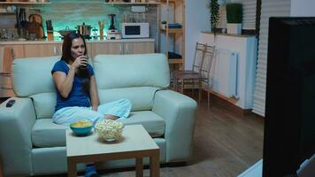 Happy woman sitting on couch watching movie on television at home. Excited, amused, lonely lady in pijamas enjoying the evening sitting on comfortable sofa in front of television eating popcorn. photo