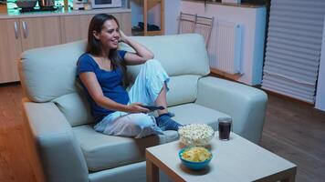 divertido mujer disfrutando gratis hora acecho televisión con palomitas de maiz y jugo. joven feliz, entusiasmado, solitario dama en pijama relajante sentado en cómodo sofá en frente de televisión sonriente y comiendo meriendas. foto