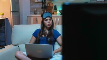 Lonely woman in pijamas and mask for sleeping on forehead sitting on couch searching information on laptop. Focused lady writing an email using internet technology late at night in living room photo