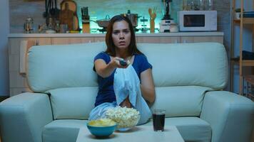 Young woman using remote controler sitting on sofa at home. Excited amused alone lady in pijamas resting with snacks and juice sitting on comfortable sofa in open space living room watching tv photo