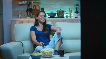 mujer riendo acecho televisión y come meriendas. joven feliz, entusiasmado, divertido, hogar solo dama disfrutando el noche sentado en cómodo sofá vestido en pijama comiendo palomitas de maiz en frente de televisión foto