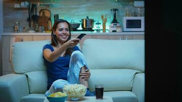 contento mujer comiendo palomitas de maiz en sofá y acecho televisión en vivo habitación a hogar. entusiasmado, divertido, solitario dama disfrutando el noche sentado en cómodo sofá vestido en pijama en frente de televisión. foto