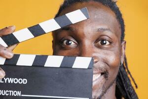 Portrait of man holding clapperboard to cut scenes in movie industry, posing on yellow background. African american man working in film making production and cinematography in studio. photo