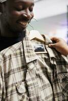 Smiling african american man holding plaid shirt while trying on outfit before making purchase in shopping mall. Clothing store customer checking apparel for wardrobe in boutique photo