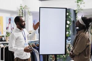 Clothing store assistant showing brand promotion on digital screen to customer. Mall worker advertising shoes new collection and offering client to check options on interactive white display photo