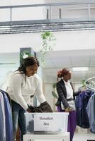 Smiling woman dropping off donations in container at charity clothing shop. African american customer putting new and used apparel in box for humanitarian aid in shopping center photo