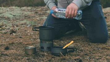 homme dans une une randonnée est cuisine et verser l'eau de le bouteille dans le pot. voyageur dans une forêt est chauffage nourriture en utilisant petit cuisinier ensemble video