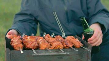 hombre es Cocinando Cerdo carne al aire libre, participación cerveza en verde botella y torneado parilla en brochetas caliente A la parrilla brocheta en el carbones picnic comida preparando. medio de cerca Disparo video