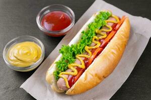 Homemade Hot Dog with mustard, ketchup, tomato and fresh salad leaves on black slate background photo