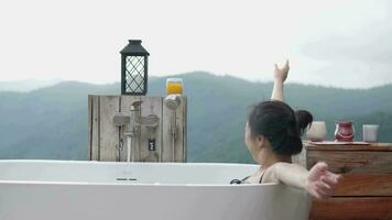 One Asian woman leisurely relaxes in a bathtub and drinks orange juice, resting in panoramic mountain range view of tropical forest, the tranquil unseen scenery natural travel resort for vacation. video