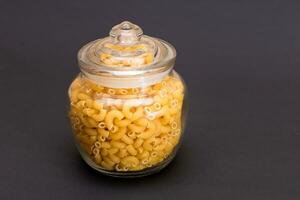 Uncooked Chifferi Rigati Pasta in Glass Jar on Black Background. Fat and Unhealthy Food. Classic Dry Macaroni. Italian Culture and Cuisine. Raw Pasta photo