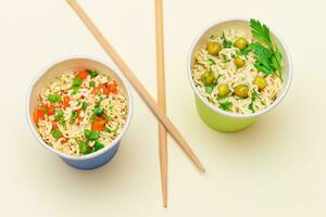 Prepared Instant Noodles with Green Pea, Carrot and Greens in Blue and Green Cups with Chopsticks on White Background. Quick Lunch or Unhealthy Fast Food photo