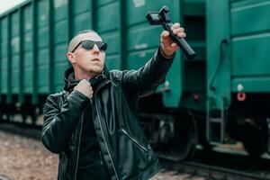 Handsome Guy in Black Clothes and Sunglasses Making Selfie or Streaming Video Using Action Camera with Gimbal Camera Stabilizer at Railway. Young Man Makes Photo Against the Cargo Wagons