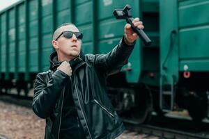 hermoso chico en negro ropa y Gafas de sol haciendo selfie o transmisión vídeo utilizando acción cámara con cardán cámara estabilizador a ferrocarril. joven hombre hace foto en contra el carga vagones