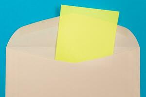 Beige Envelope with Blank Yellow Sheet of Paper Inside, Lying on Blue Background Mock Up with Copy Space. Receiving Mail or Notice, Sending Postcard Top View, Flat Lay photo