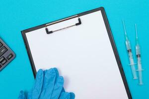 Vaccination, Immunology or Revaccination Concept Two Medical Syringe Lying on Blue Table in Doctor's Office in a Hospital or Clinic. Black Clipboard with Sheet of Paper Mock Up with Copy Space photo